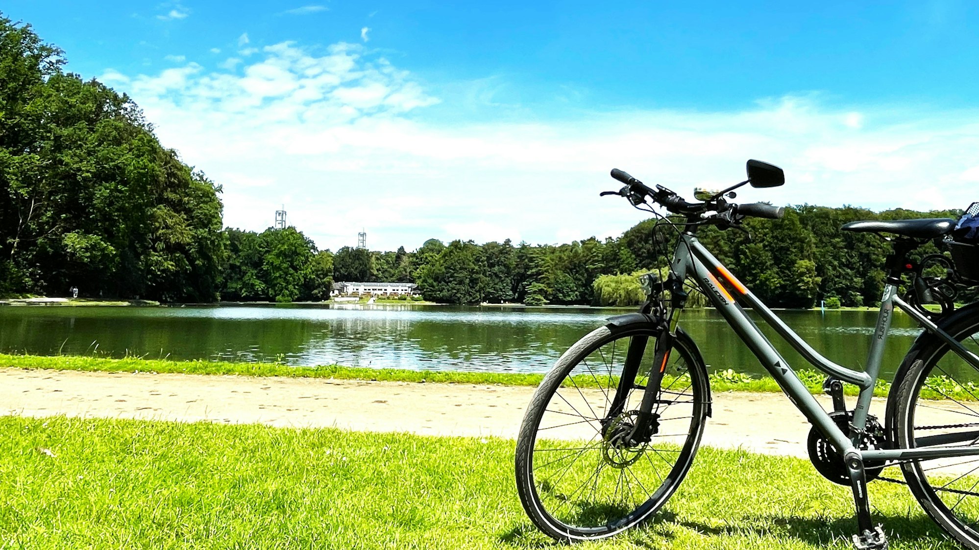 Ein Fahrrad steht auf der Wiese am Adenauerweiher in Köln. In der Ferne sind der Club Astoria und die Stützen vom Rhein Energie Stadion zu sehen.