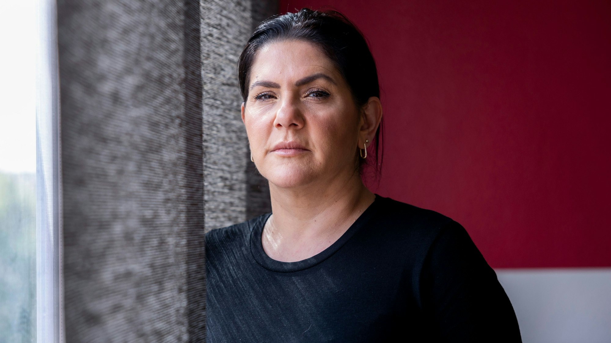 Mariam Claren hat schwarze Haare. Sie trägt ein schwarzes Top. Sie steht am Fenster und schaut in die Kamera.