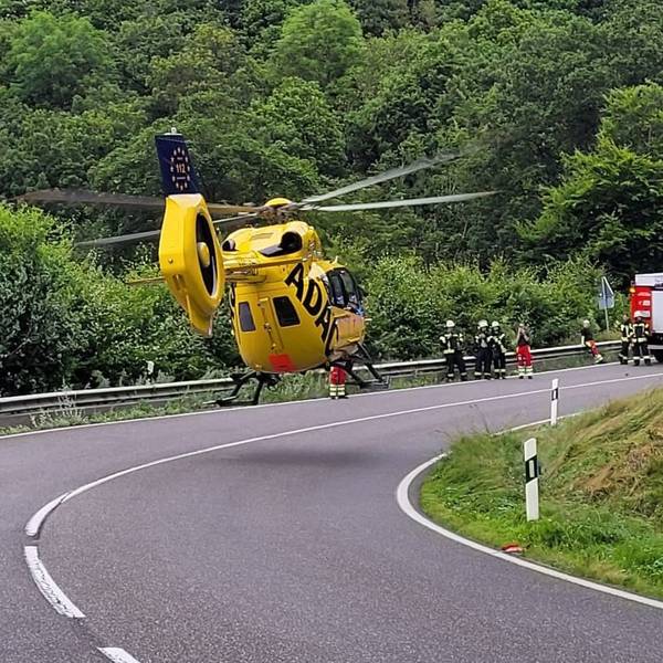 Wenige Meter über der Straße ist gelber ADAC-Rettungshubschrauber zu sehen. Im Hintergrund stehen Einsatzkräfte und ein Feuerwehrwagen.