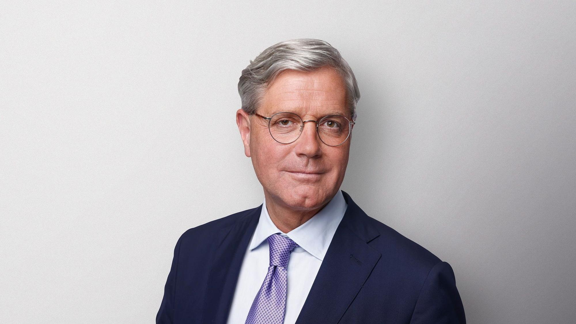 Ein Mann, der Anzug und Brille trägt, steht vor einem neutralen Hintergrund.