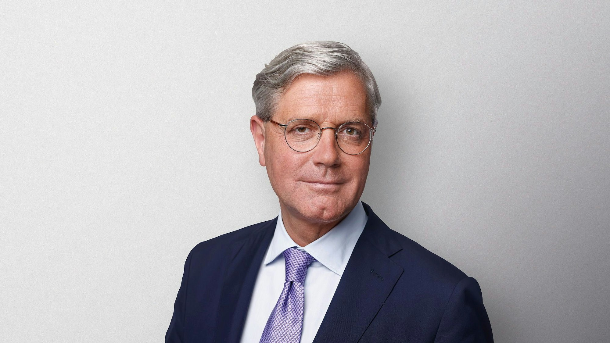 Ein Mann, der Anzug und Brille trägt, steht vor einem neutralen Hintergrund.