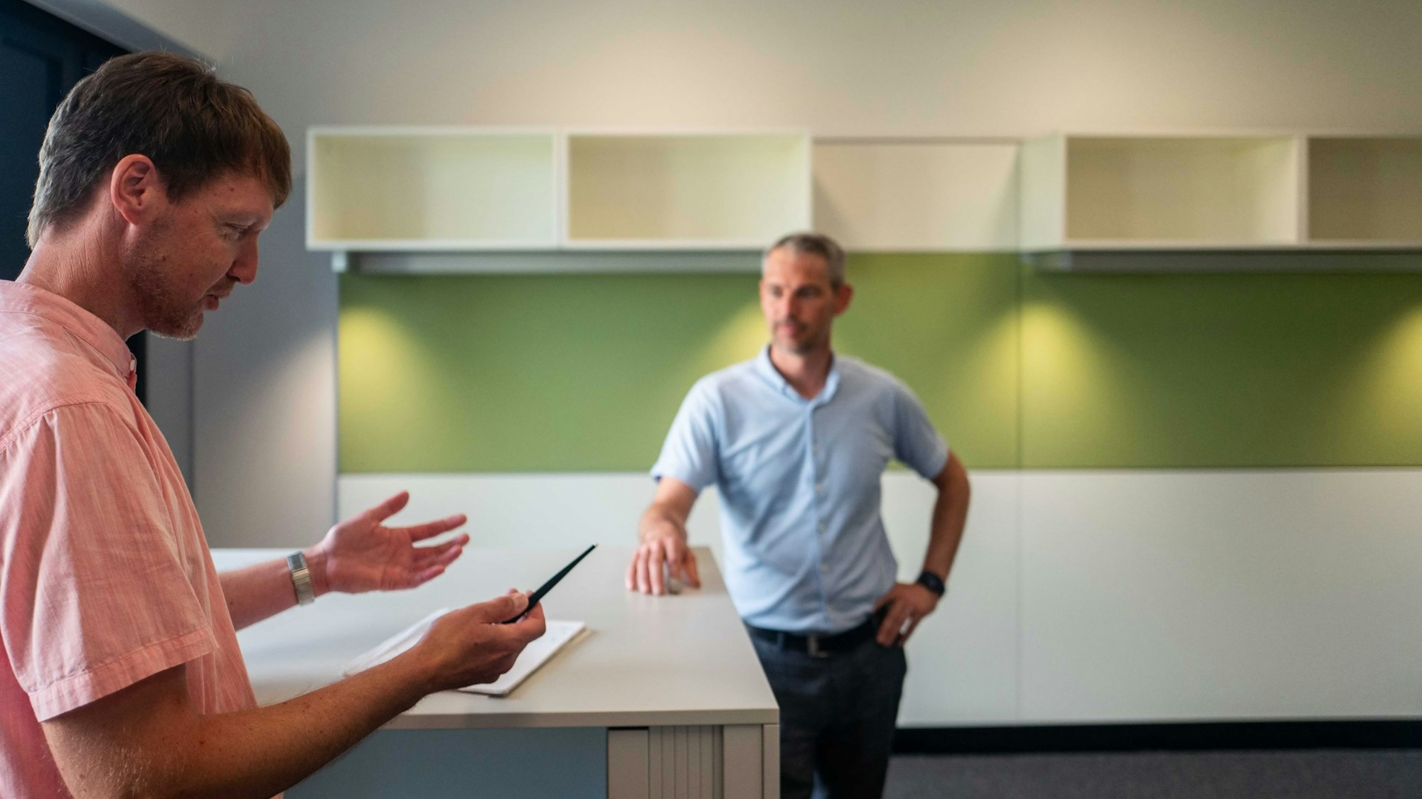 Das Bild zeigt Carsten Opitz und Guido Schmitz. Opitz steht an einem Schreibtisch und erklärt Schmitz die Aufteilung des Büros.