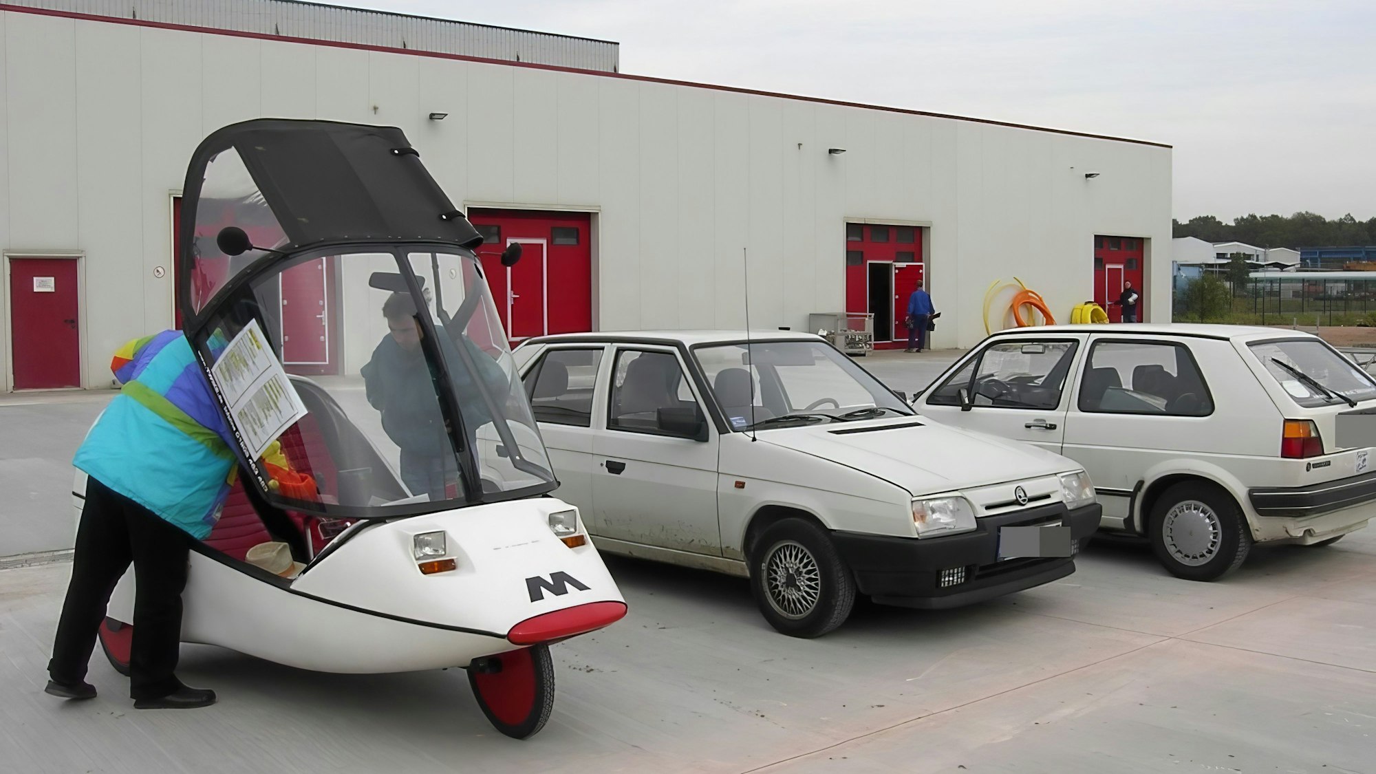 Die drei Fahrzeuge stehen nebeneinander. Zwei Besucher schauen in das Innere des Dreiradwagens, dessen Verdeck hochgehoben worden ist.