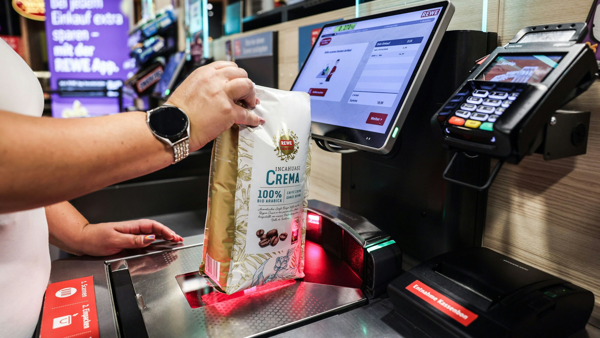 Eine Frau scannt in einer Rewe-Filiale ein Produkt an einer Selbstbedienungskasse.
