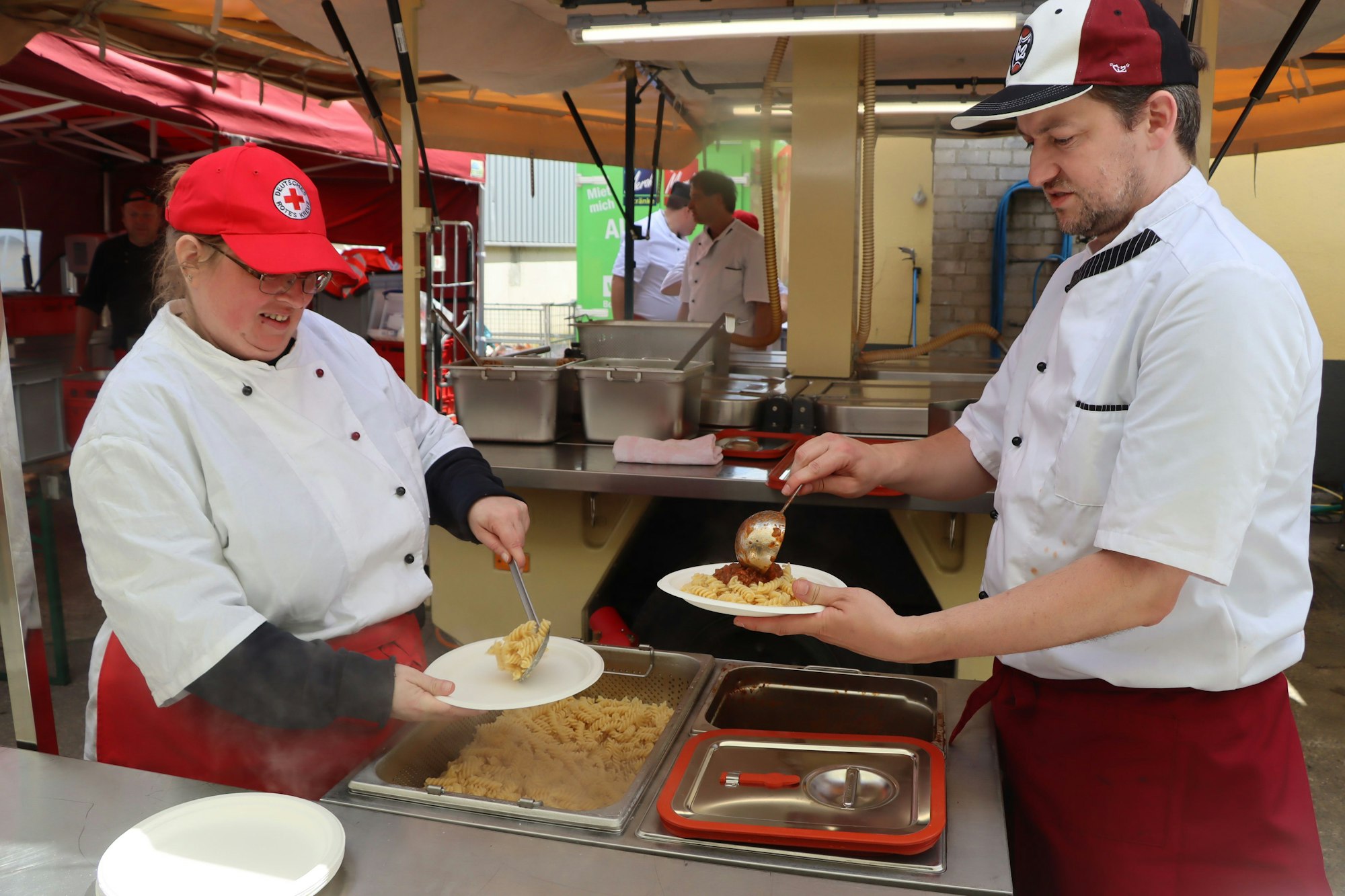 Das Bild zeigt zwei Personen in einer Großküche, die Essen ausgeben.