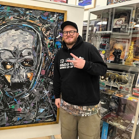 Der Langenfelder Künstler und Store-Betreiber Ivan Beslic steht vor dem von ihm gezeichneten Gemälde des Rappers Sido in seiner früheren Erscheinung mit Maske. Beslic zeigt mit zwei Fingern das Peace-Zeichen, hinter ihm sind Ausstellungsstücke hinter einer Vitrine zu sehen.