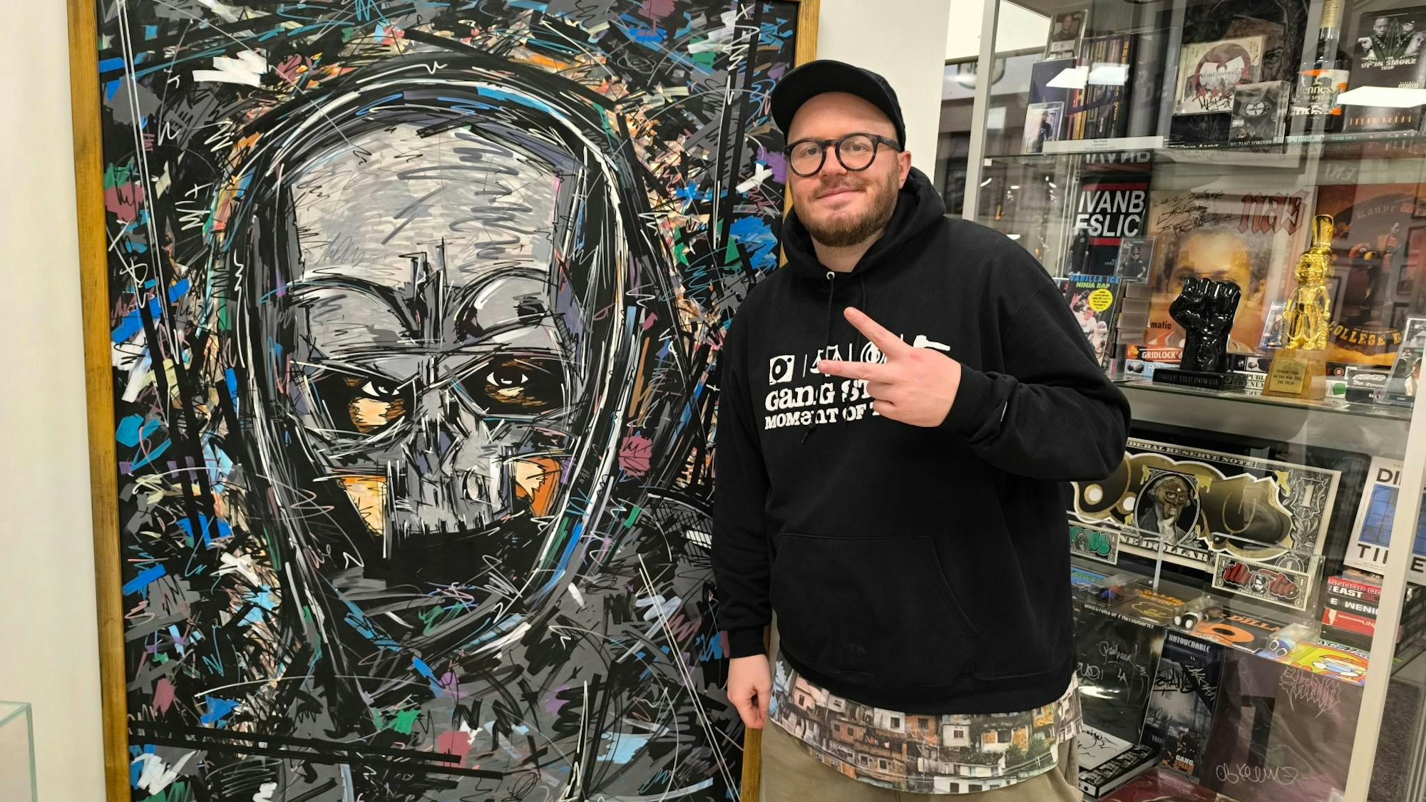 Der Langenfelder Künstler und Store-Betreiber Ivan Beslic steht vor dem von ihm gezeichneten Gemälde des Rappers Sido in seiner früheren Erscheinung mit Maske. Beslic zeigt mit zwei Fingern das Peace-Zeichen, hinter ihm sind Ausstellungsstücke hinter einer Vitrine zu sehen.