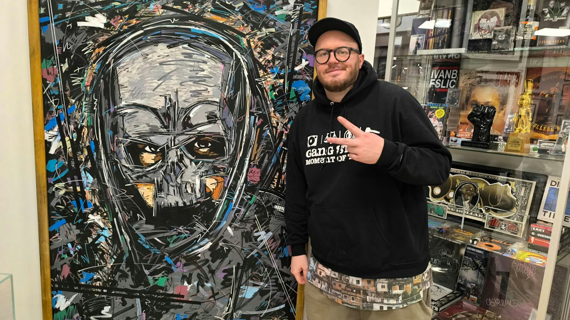 Der Langenfelder Künstler und Store-Betreiber Ivan Beslic steht vor dem von ihm gezeichneten Gemälde des Rappers Sido in seiner früheren Erscheinung mit Maske. Beslic zeigt mit zwei Fingern das Peace-Zeichen, hinter ihm sind Ausstellungsstücke hinter einer Vitrine zu sehen.