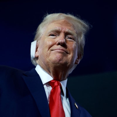 Vor dunklem Hintergrund hebt sich das lächelnde Gesicht Donald Trumps ab. Über dem rechten Ohr trägt er einen Verband, Zeugnis der Verletzung durch ein Attentat während einer Wahlkampfveranstaltung am 13. Juli.