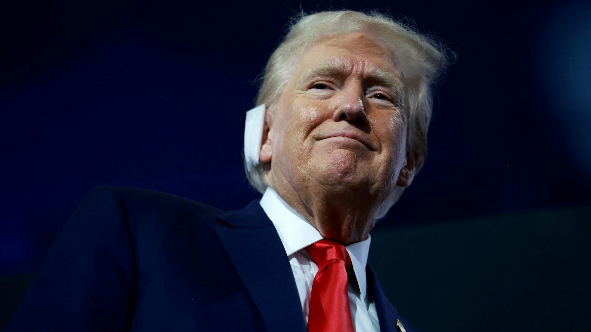 Vor dunklem Hintergrund hebt sich das lächelnde Gesicht Donald Trumps ab. Über dem rechten Ohr trägt er einen Verband, Zeugnis der Verletzung durch ein Attentat während einer Wahlkampfveranstaltung am 13. Juli.