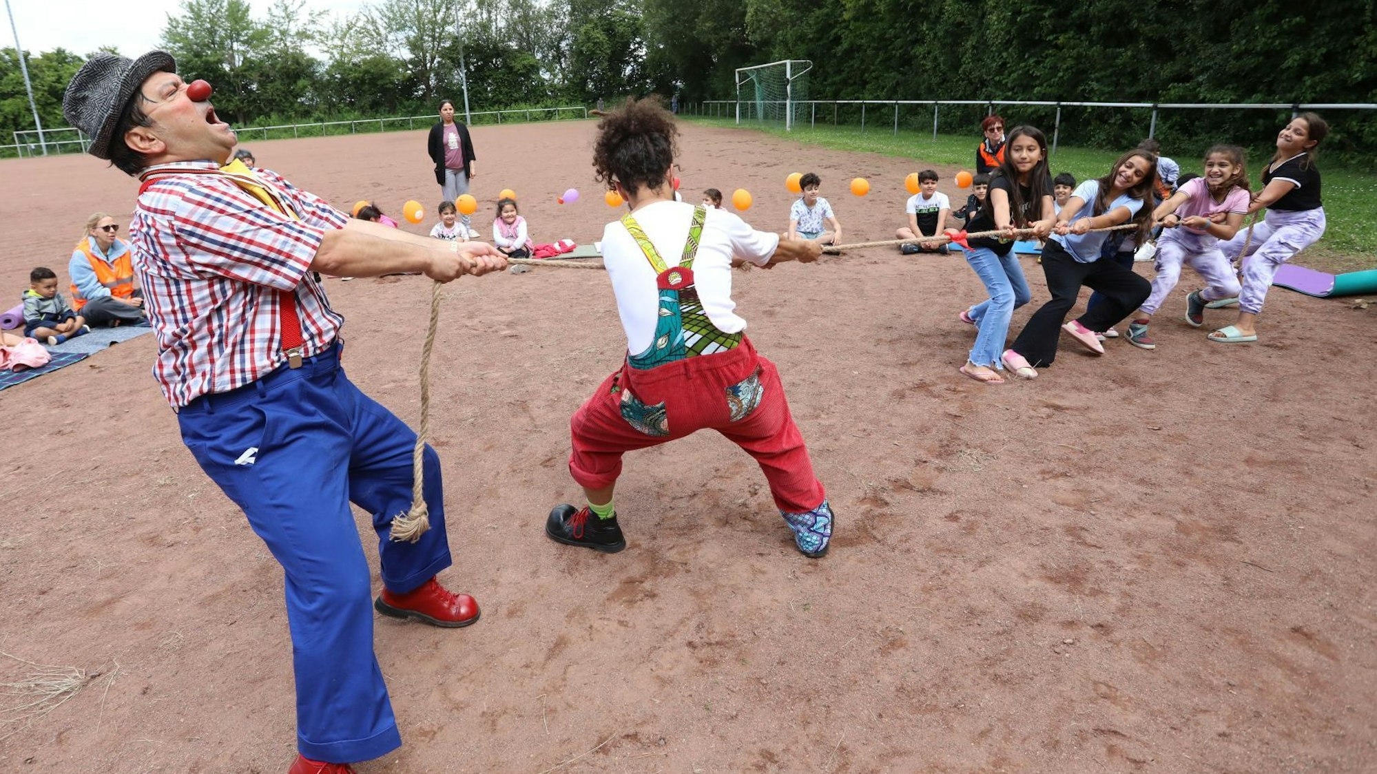 Zwei bunt gekleidete Clowns wetteifern beim Seil ziehen gegen eine Gruppe Kinder.