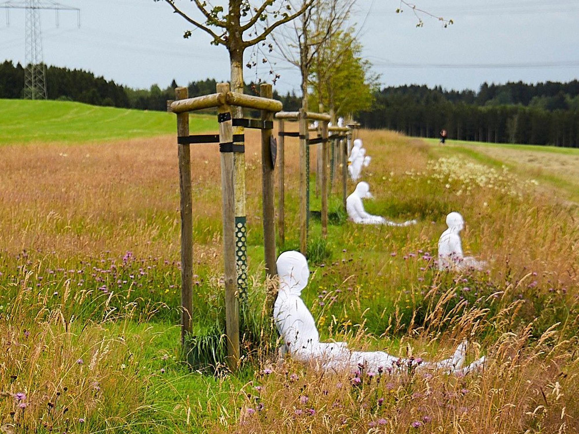 In weiße Anzüge gekleidete Menschen sitzen vor den Bäumen der zentralen NRW-Gedenkstätte für die Opfer der Flutkatastrophe. Sie symbolisieren die gestorbenen Menschen.