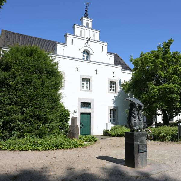 Das alte Gebäude mit weißem Anstrich ist von außen zu sehen. Davor steht ein Denkmal.