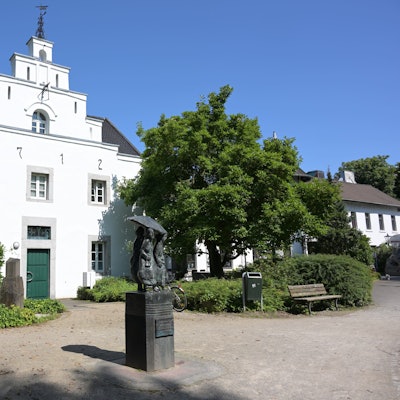 Das alte Gebäude mit weißem Anstrich ist von außen zu sehen. Davor steht ein Denkmal.