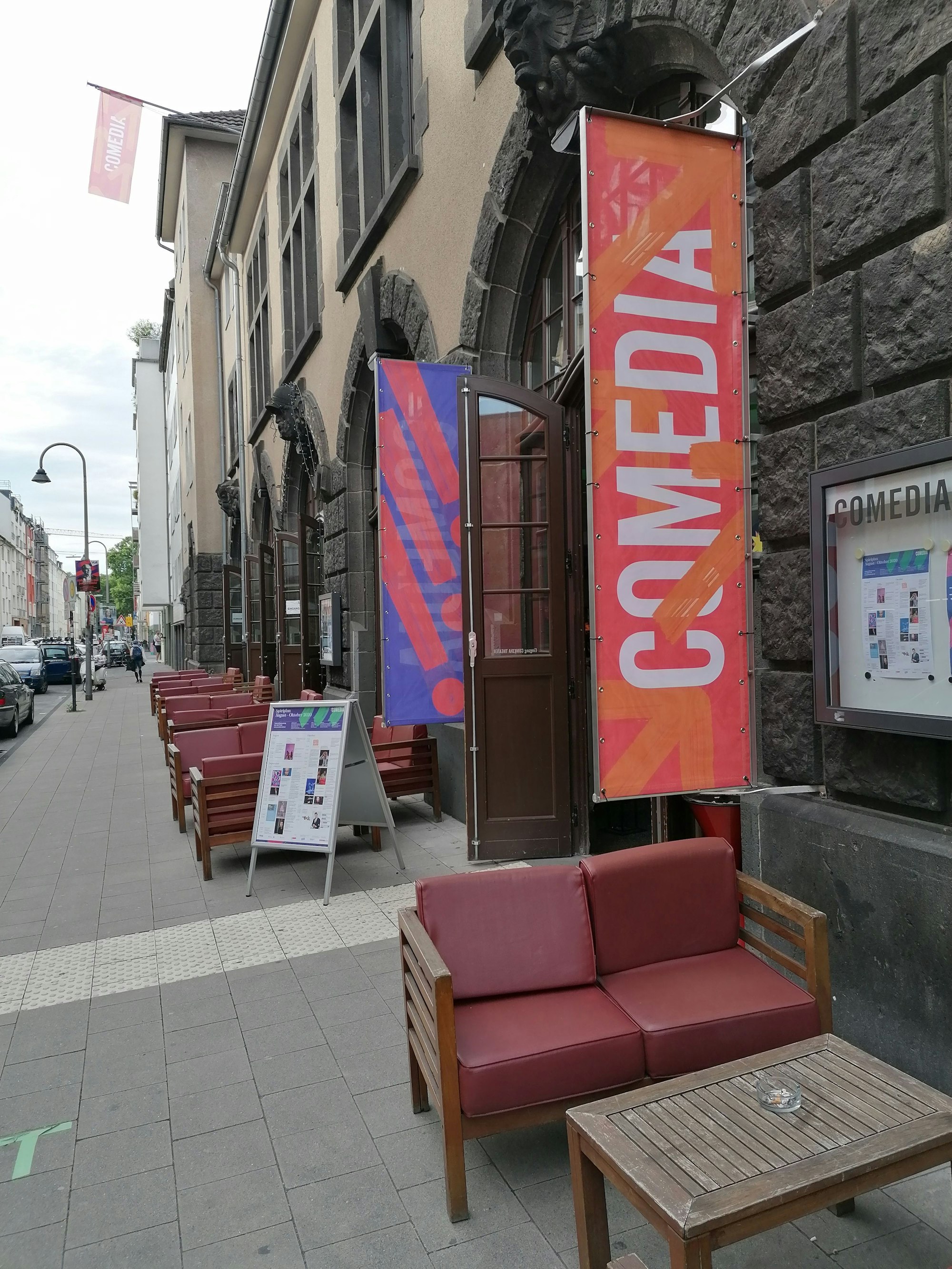 Auf dem Bild ist das Gebäude des Comedia Theaters zu sehen, davor auf dem Bürgersteig ein paar Sessel an Tischen. Am Eingang des Gebäudes ist ein großes rotes Banner mit der Aufschrift „COMEDIA“ zu sehen.