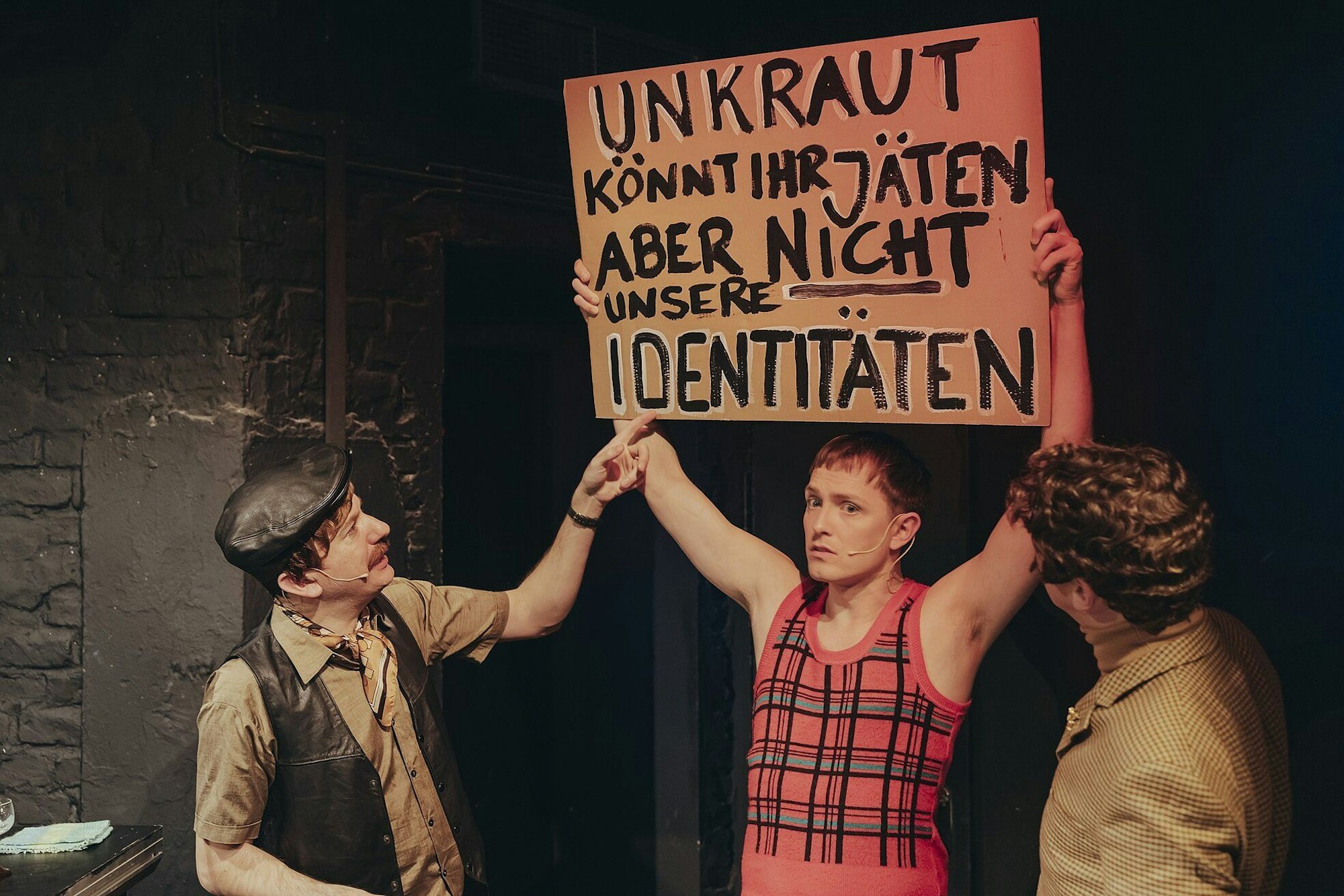 Ein Mann hält im Protest ein Schild hoch, auf dem Steht: „Unkraut könnt ihr jäten, aber nicht unsere Identitäten“. Er trägt ein ärmelloses rotes Oberteil mit Quer- und Längsstreifen. Zwei andere Männer schauen sich das Schild an, einer zeigt darauf.
