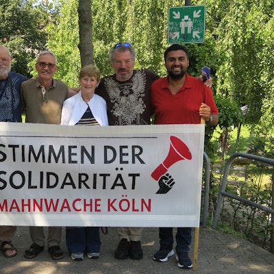 Veranstalter des „Festival der Solidarität“ in Köln-Ehrenfeld stehen hinter einem Banner mit der Aufschrift „Stimmen der Solidarität – Mahnwache Köln“.
