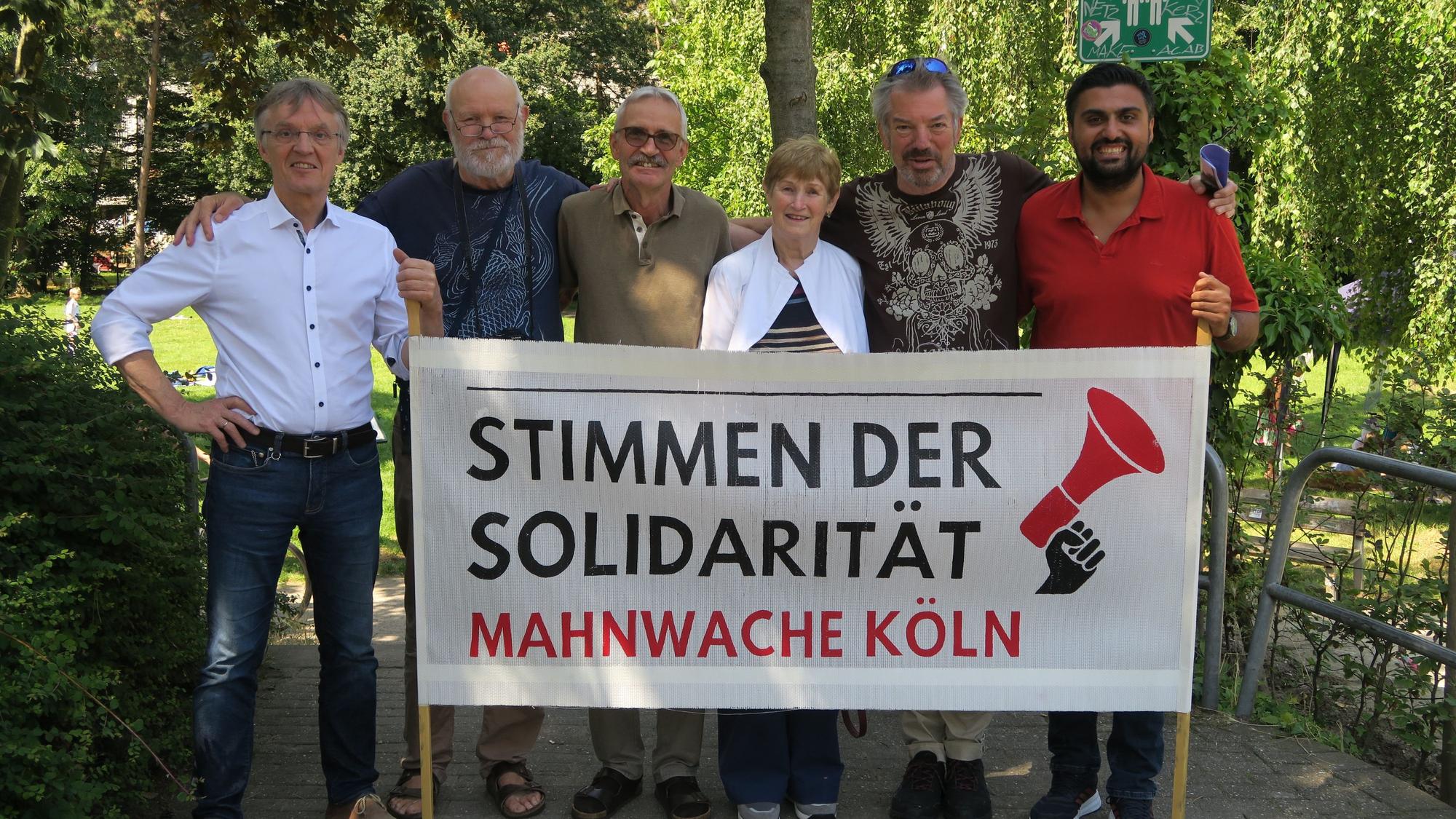 Mitglieder des Vereins „Stimmen der Solidarität – Mahnwache Köln e. V.“ stehen hinter einem Banner
