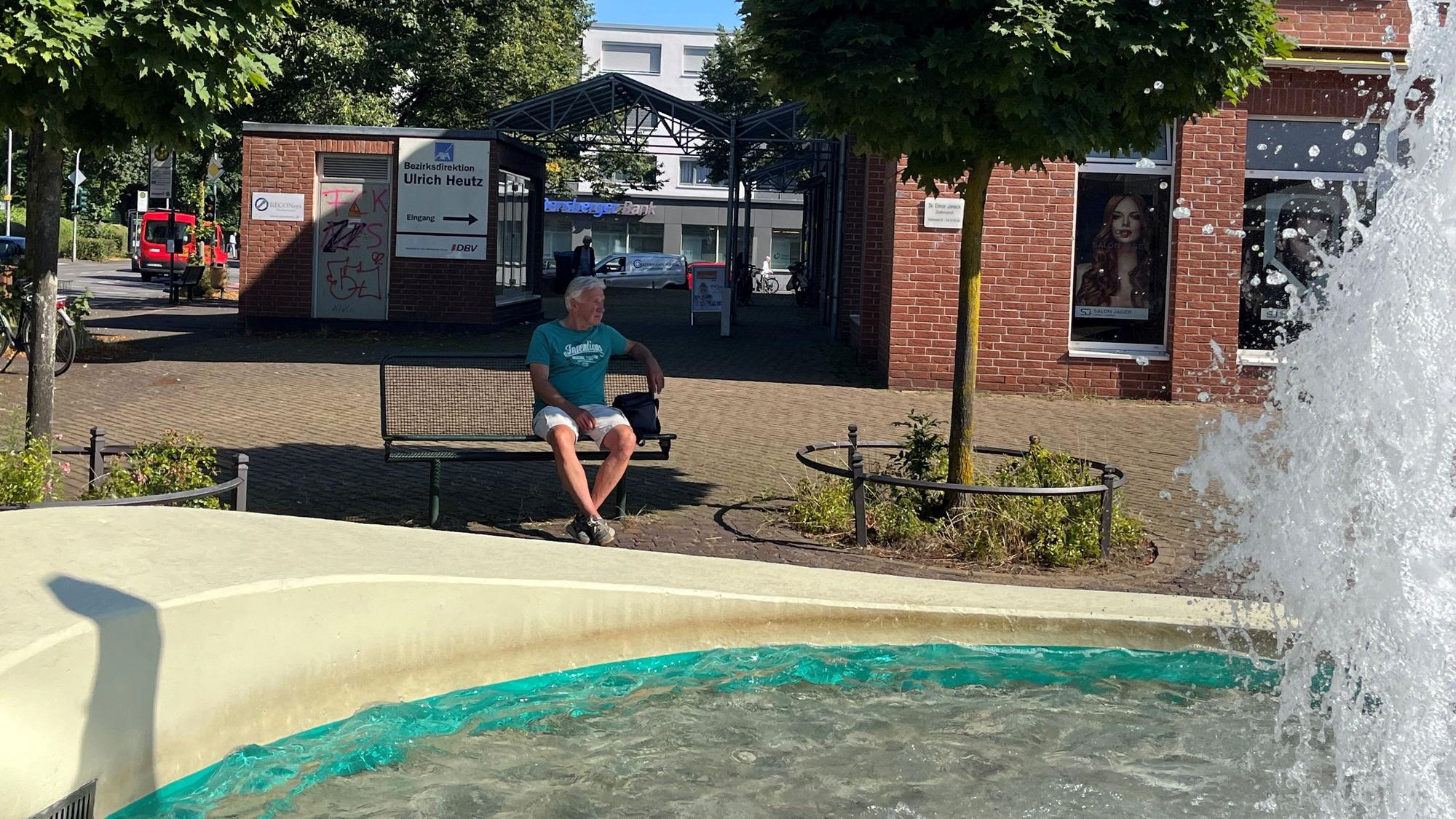 Rechts sieht man die Wasserfontäne. Der Kölner sitzt auf einer Bank.