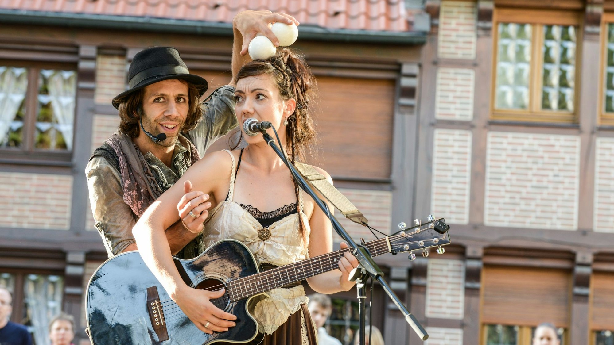 Ein Mann und eine Frau in historischen Kostümen. Er hält Jonglierbälle über ihren Kopf, die Frau spielt Gitarre.