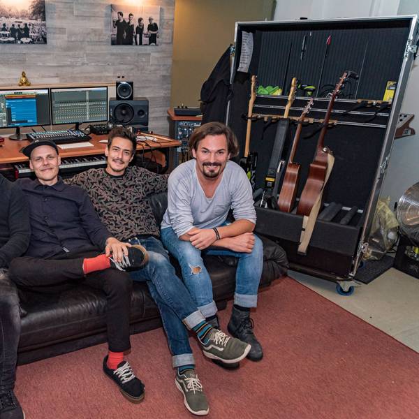 Vier Männer sitzen nebeneinander auf einem Sofa, hinter ihnen sind ein Computer und Musikboxen sowie Gitarren.