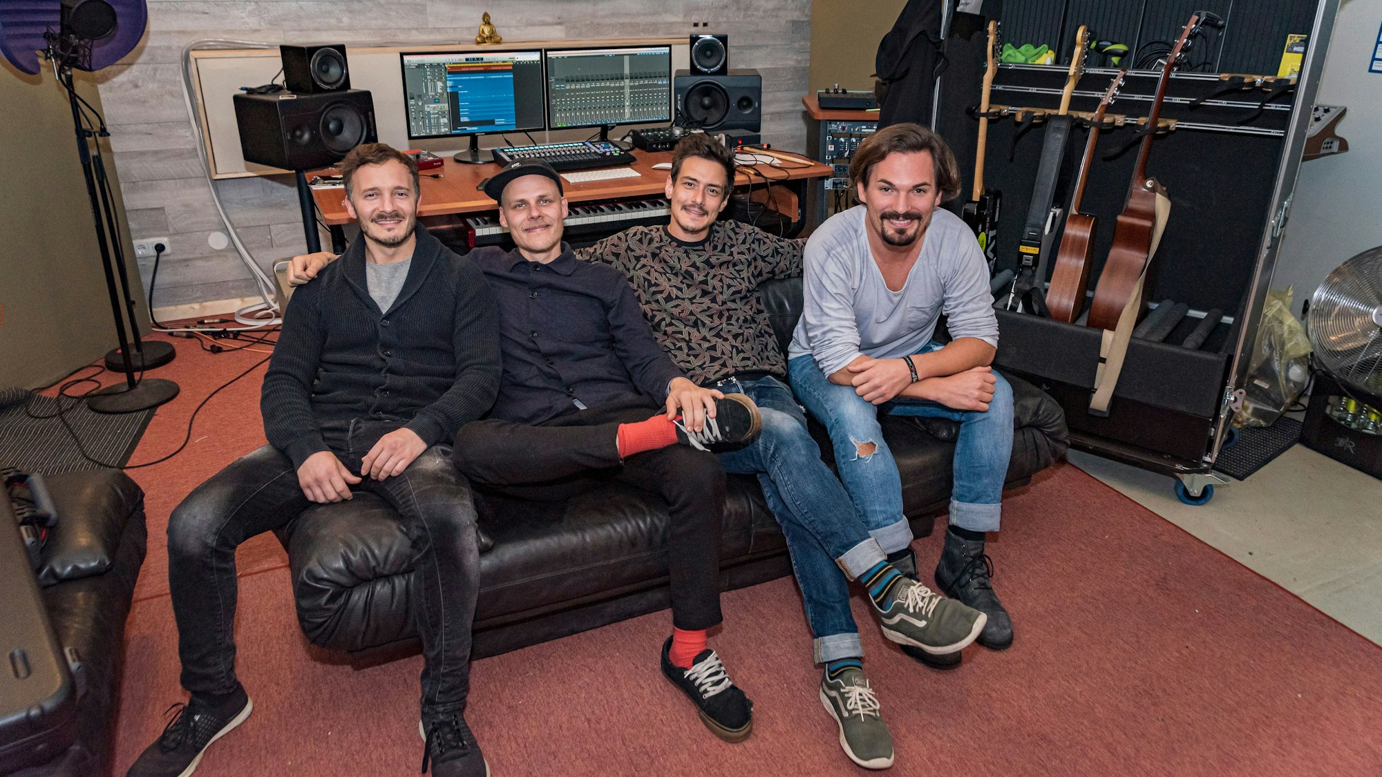 Vier Männer sitzen nebeneinander auf einem Sofa, hinter ihnen sind ein Computer und Musikboxen sowie Gitarren.