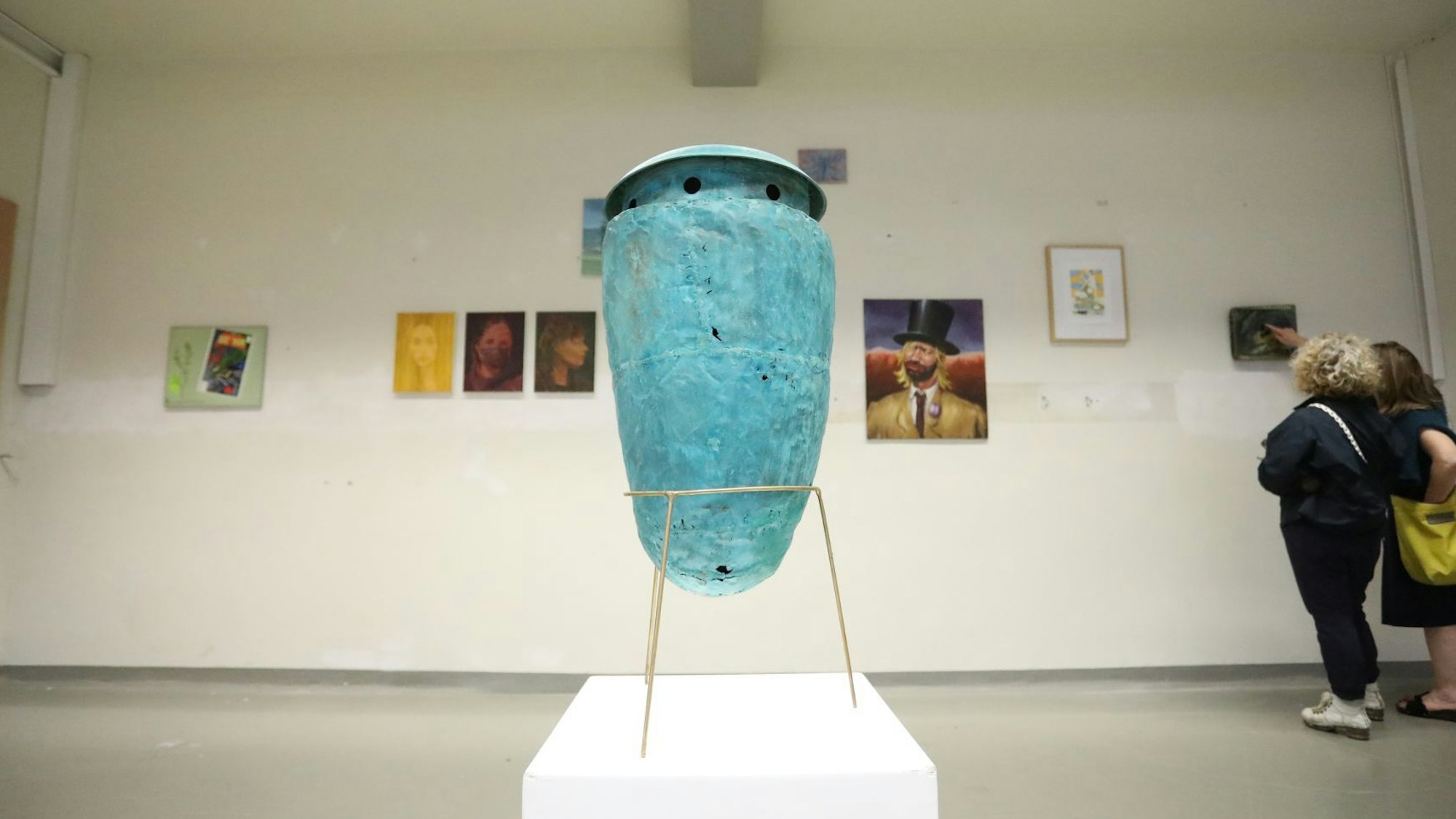 Eine an eine Vase erinnernde Skulptur steht auf einem weißen Sockel, im Hintergrund hängen Kunstwerke an der Wand-