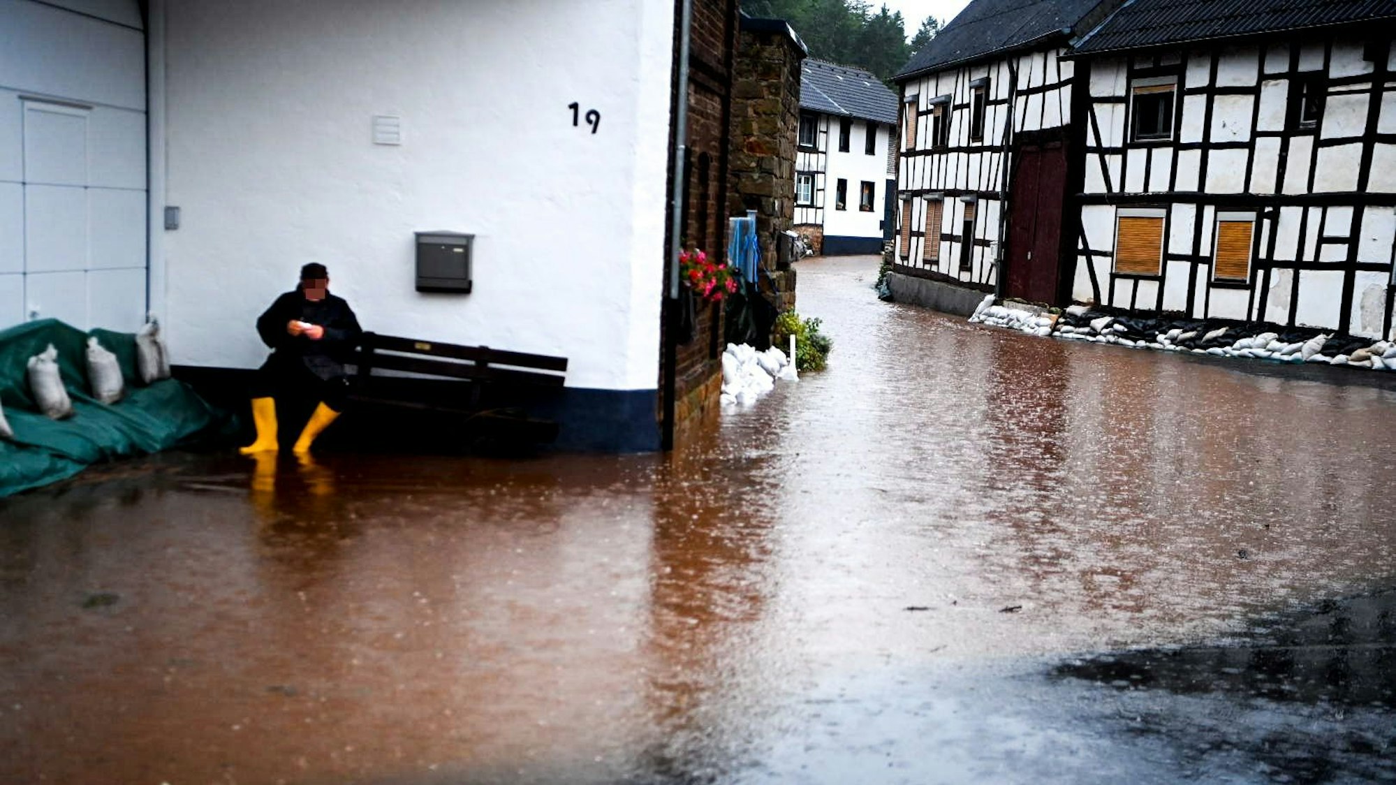 Ein Anwohner im schon leicht überschwemmten Ort Gilsdorf sitzt auf einer Bank und ruht sich aus. Er trägt gelbe Gummistiefel.