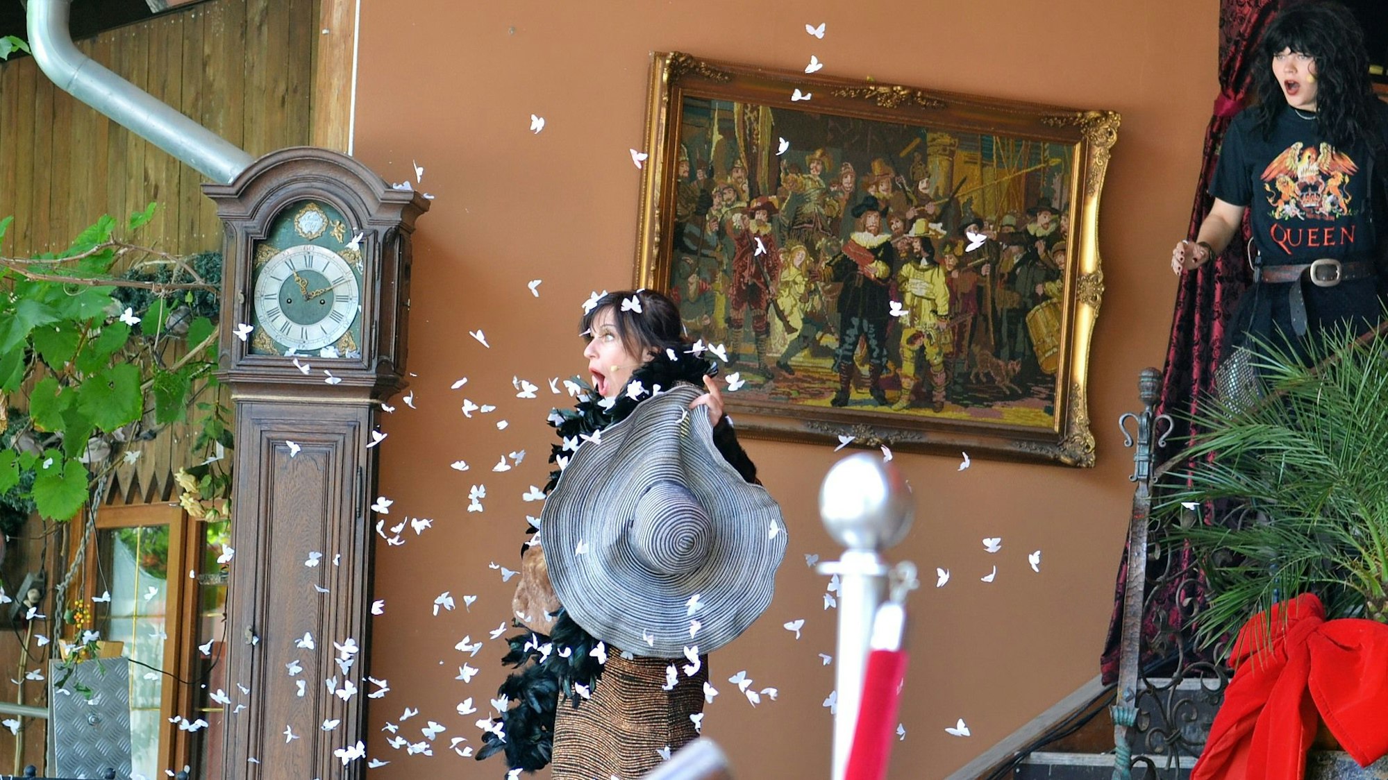 Eine Frau mit einem großen Hut in der Hand hält vor lauter Schreck auf einer Treppe inne. Dutzende weiße Falter umkreisen sie. Im Hintergrund eine Standuhr und ein Ölgemälde.