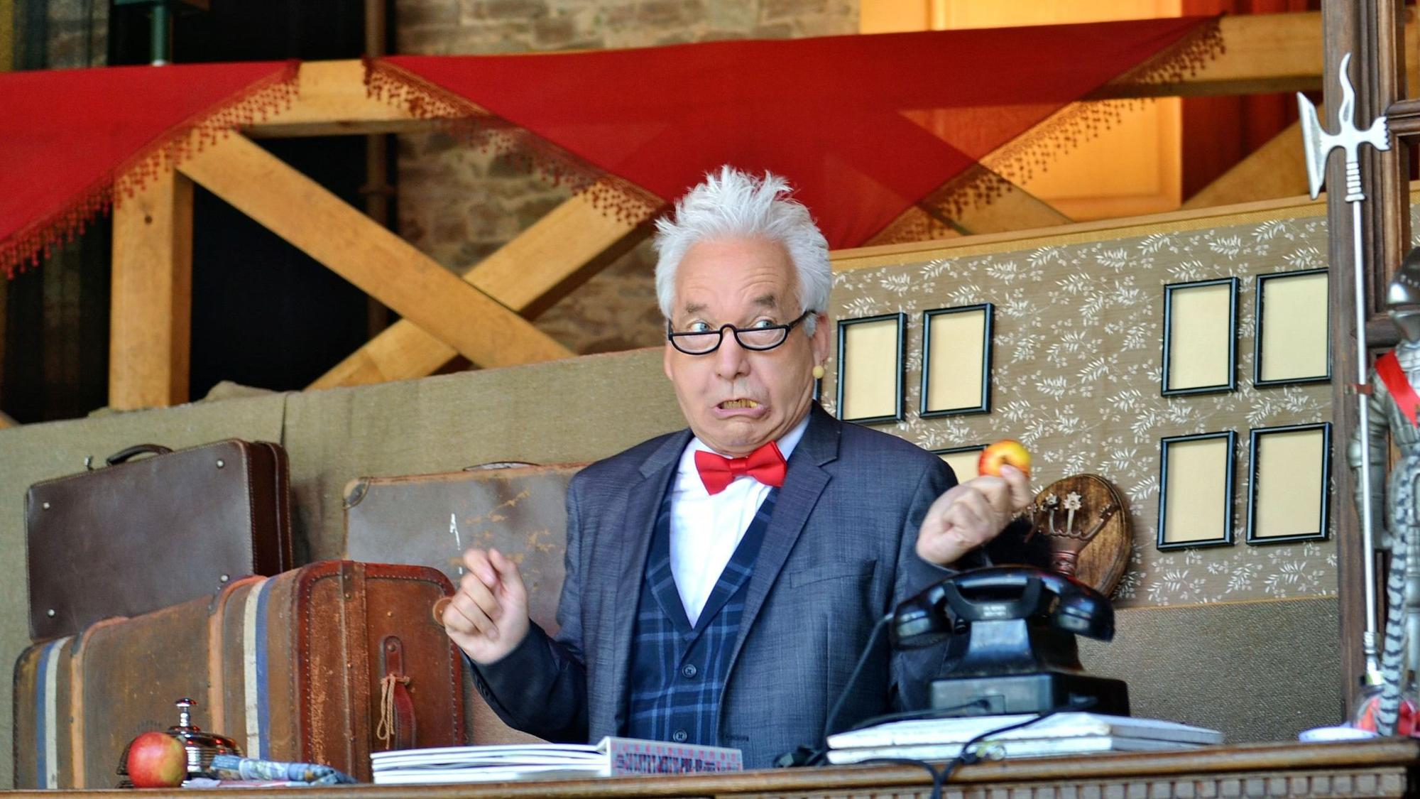Claus Vinçon sitzt als Freiherr von und zu Fronzenberg an der Hotelrezeption.
