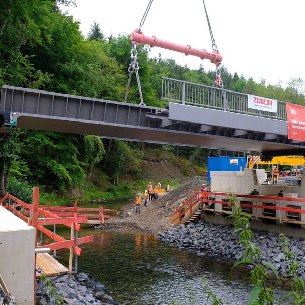 Mithilfe eines Mobilkrans wird eine Stahlbrücke auf die Widerlager gehoben. Darunter fließt die Urft in ihrem Flussbett.