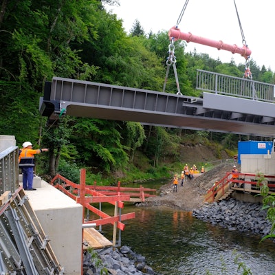 Mithilfe eines Mobilkrans wird eine Stahlbrücke auf die Widerlager gehoben. Darunter fließt die Urft in ihrem Flussbett.