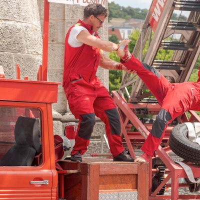 Ein Mann und eine Frau in Feuerwehranzügen machen akrobatische Kunststücke auf einem Feuerwehrauto.