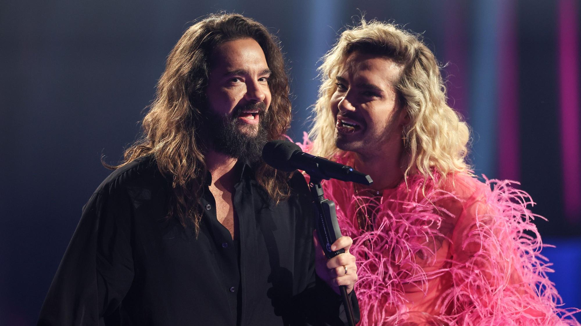 Tom und Bill Kaulitz stehen vor einem Mikrofon und lächeln.