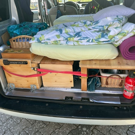 Das Bild zeigt den hinteren Bereich eines Renault Kangoo. Mit Holzaufbauten ist eine Schlaffläche samt Stauraum entstanden. Darauf liegt eine Matratze.