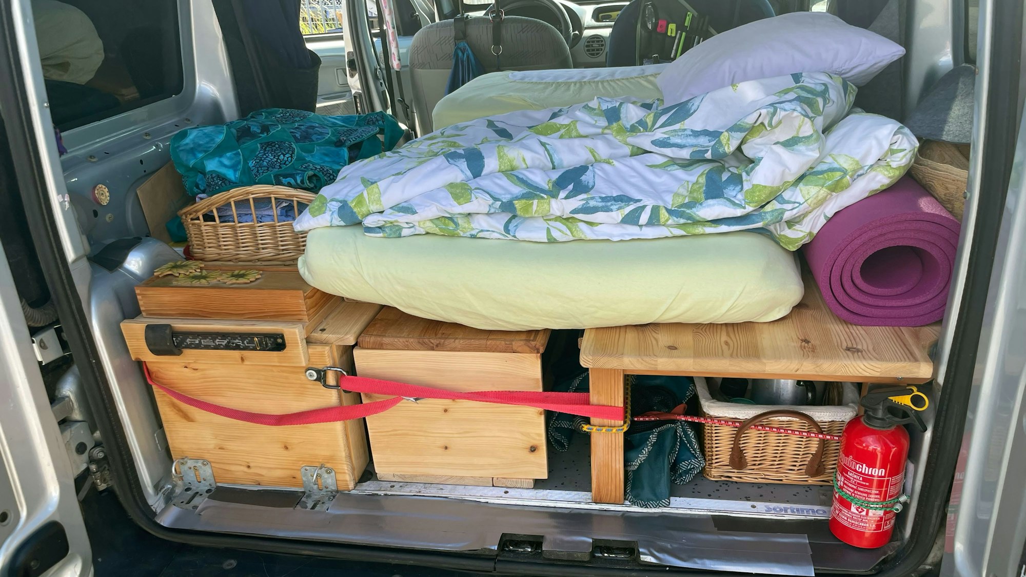 Das Bild zeigt den hinteren Bereich eines Renault Kangoo. Mit Holzaufbauten ist eine Schlaffläche samt Stauraum entstanden. Darauf liegt eine Matratze.