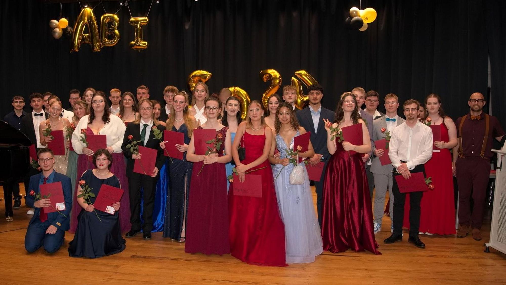 Der Abiturjahrgang 2024 der Gesamtschule Mechernich steht in Abendgaderobe auf einer mit goldenen Luftballons dekorierten Bühne.