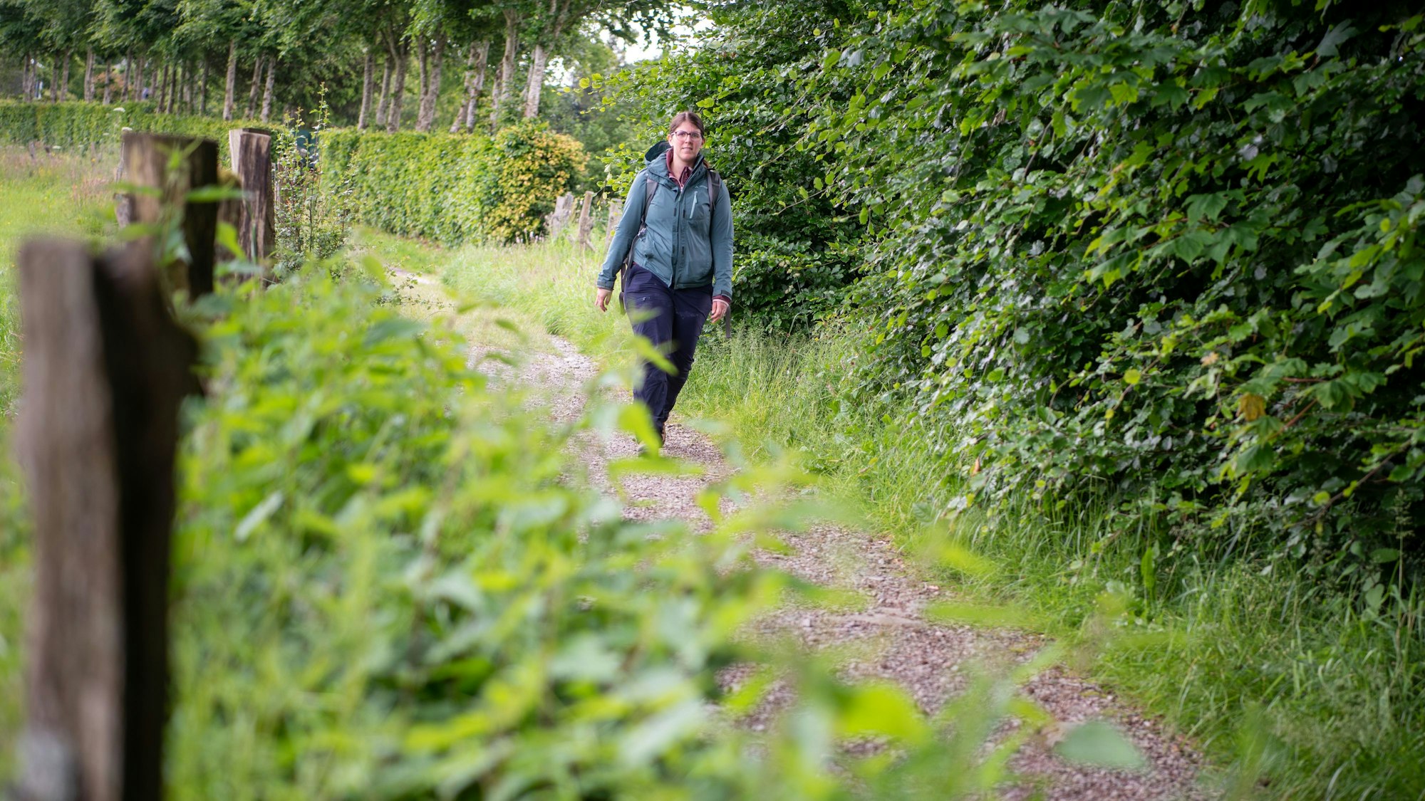 Eine Frau in grüner Jacke und dunkler Hose läuft über einen Wanderweg, rundherum sind Wiesen, Hecken, und Bäume zu sehen.