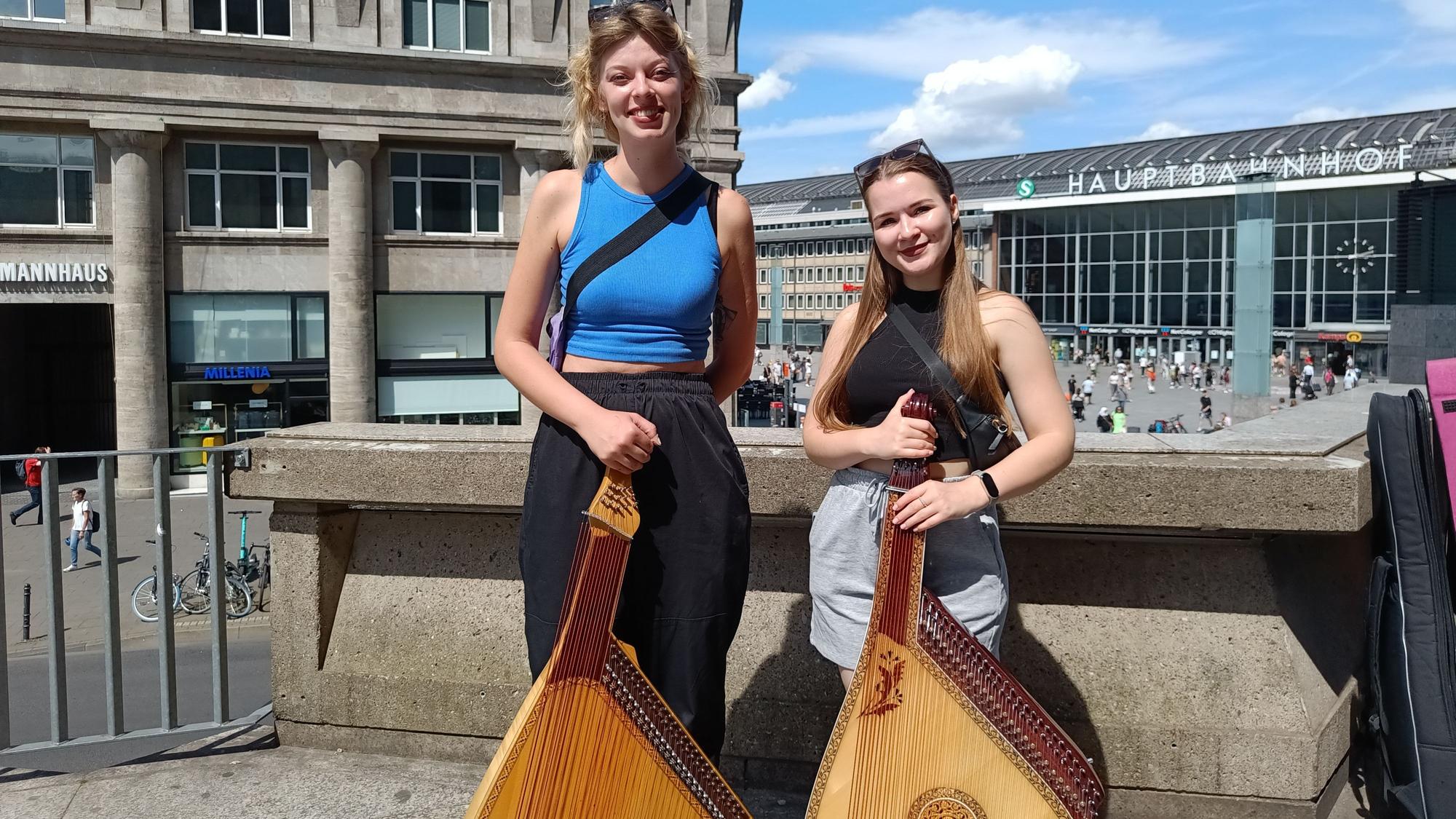 Zwei Frauen mit gitarrenähnlichen Musikinstrumenten stehen im Schatten des Kölner Doms.