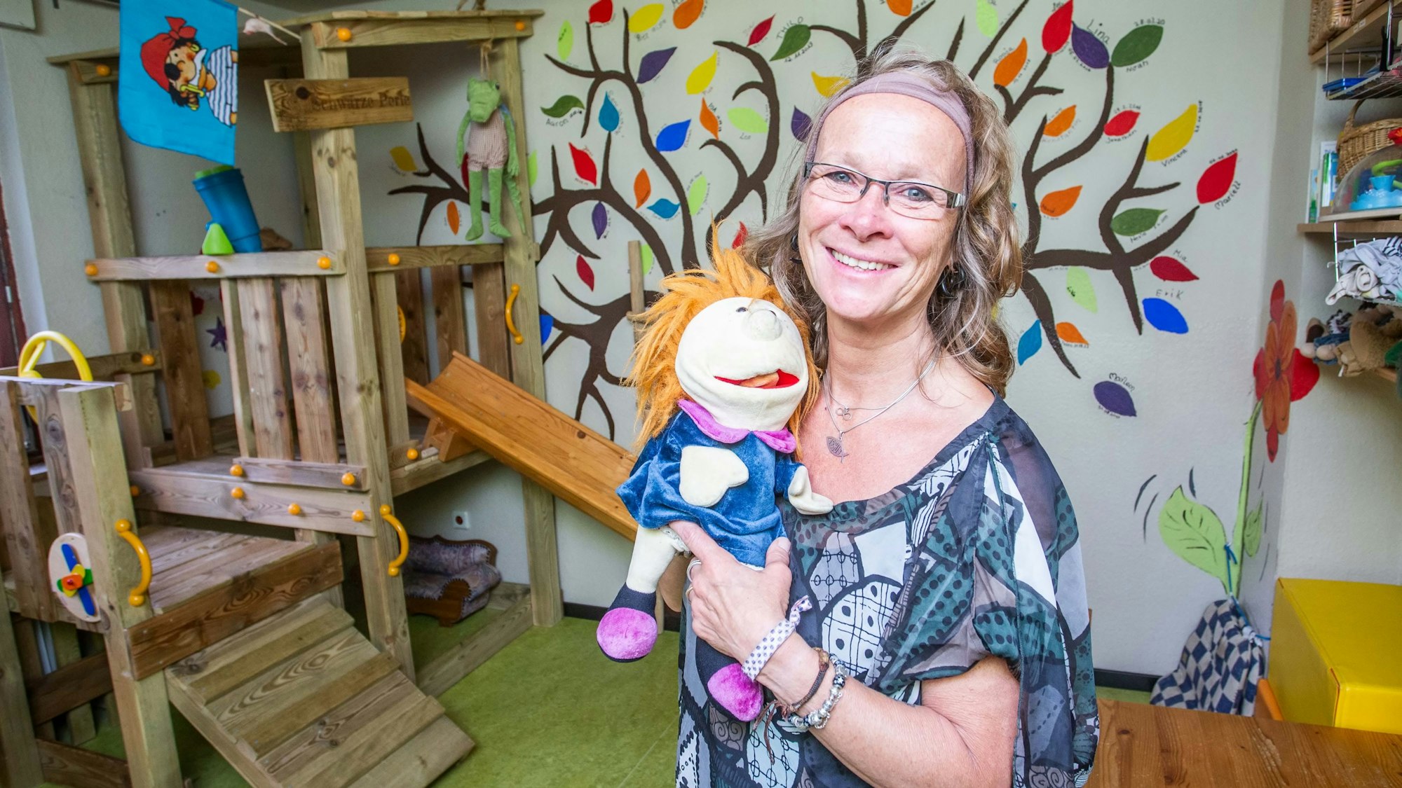 Silke Kappenberg steht mit einer Handpuppe in der Hand in einem Kinderzimmer. Im Hintergrund ist ein großer Baum zu sehen, der mit bunten Farben an die Wand gemalt wurde. Auf jedem Blatt steht der Name eines ihrer Tageskinder.