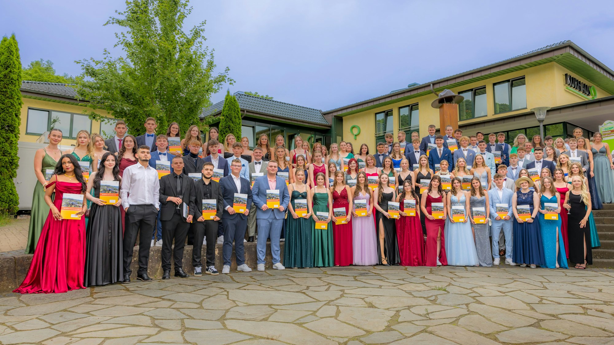 Der Abiturjahrgang der Clara-Fey-Schule steht in festlicher Garderobe vor dem gelben Kurhaus in Gemünd und hält seine Zeugnisse in der Hand.