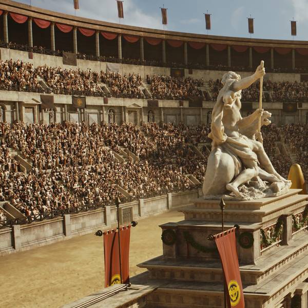 Auf dem Bild ist eine große Arena zu sehen. Es findet ein Wagenrennen statt. Die Wagen fahren um ein Monument inmitten der Arena herum, darauf thront eine Statue.