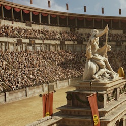 Auf dem Bild ist eine große Arena zu sehen. Es findet ein Wagenrennen statt. Die Wagen fahren um ein Monument inmitten der Arena herum, darauf thront eine Statue.