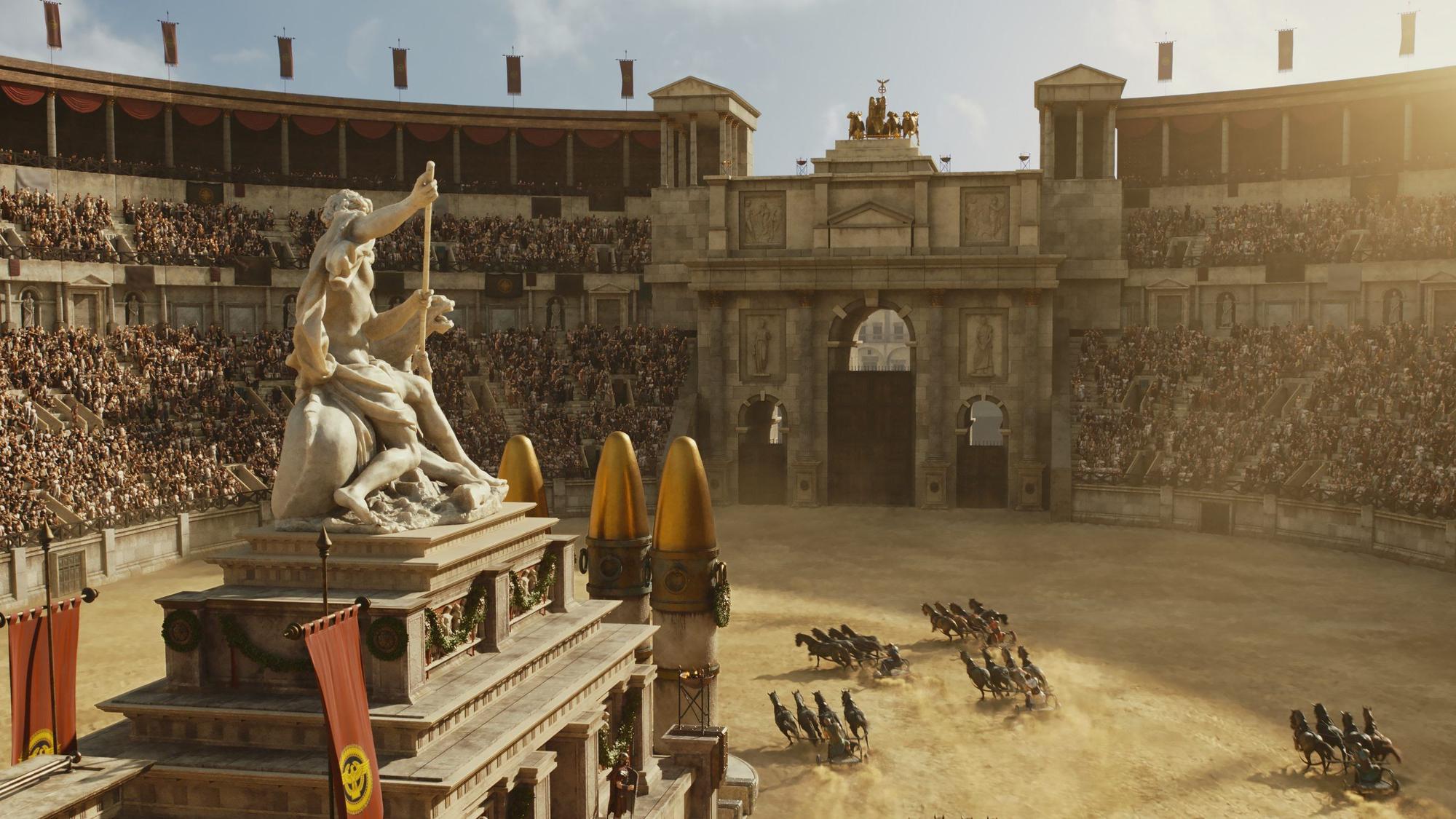 Auf dem Bild ist eine große Arena zu sehen. Es findet ein Wagenrennen statt. Die Wagen fahren um ein Monument inmitten der Arena herum, darauf thront eine Statue.