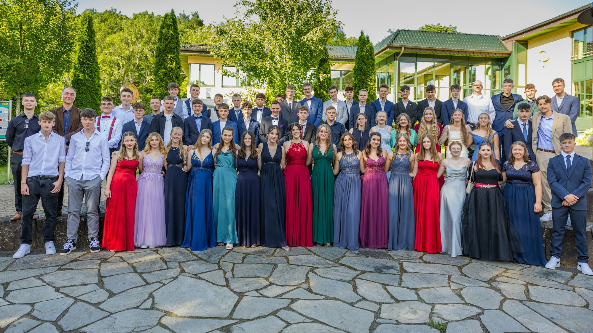 Der Abschlussjahrgang der Clara-Fey-Realschule steht in Abendgarderobe auf einem Platz. Im Hintergrund ist das gelbe Gebäude des Kurhauses in Gemünd und Wald zu erkennen.