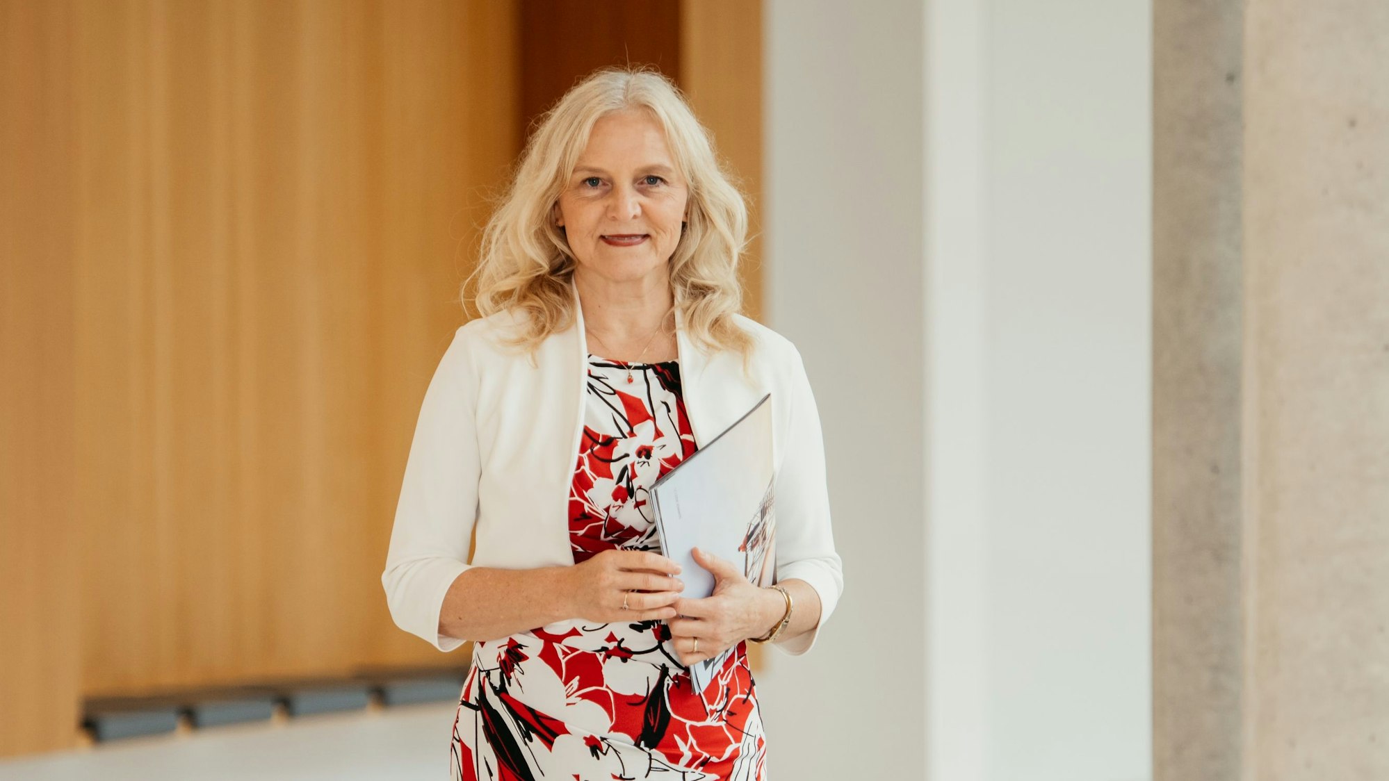 Eine Frau lächelt in die Kamera, in einer Hand hält sie Unterlagen.