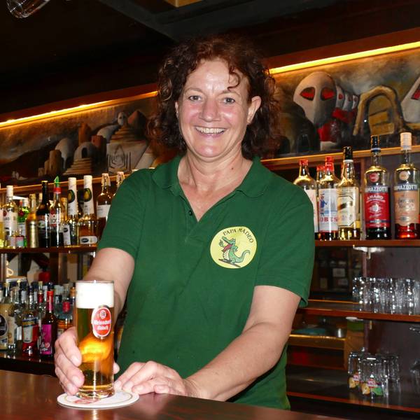 Frau in grünem T-Shirt steht hinter der Theke und stellt ein gefülltes Glas mit Kölsch auf den Tresen.