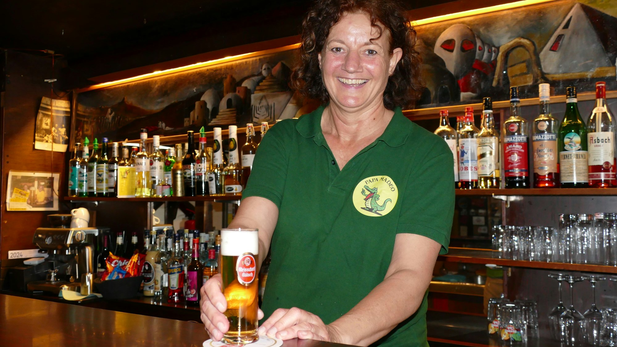 Frau in grünem T-Shirt steht hinter der Theke und stellt ein gefülltes Glas mit Kölsch auf den Tresen.