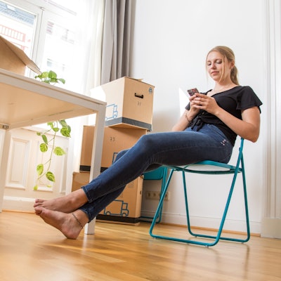 Eine Frau sitzt zwischen Umzugskartons in einer Wohnung auf einem Klappstuhl.