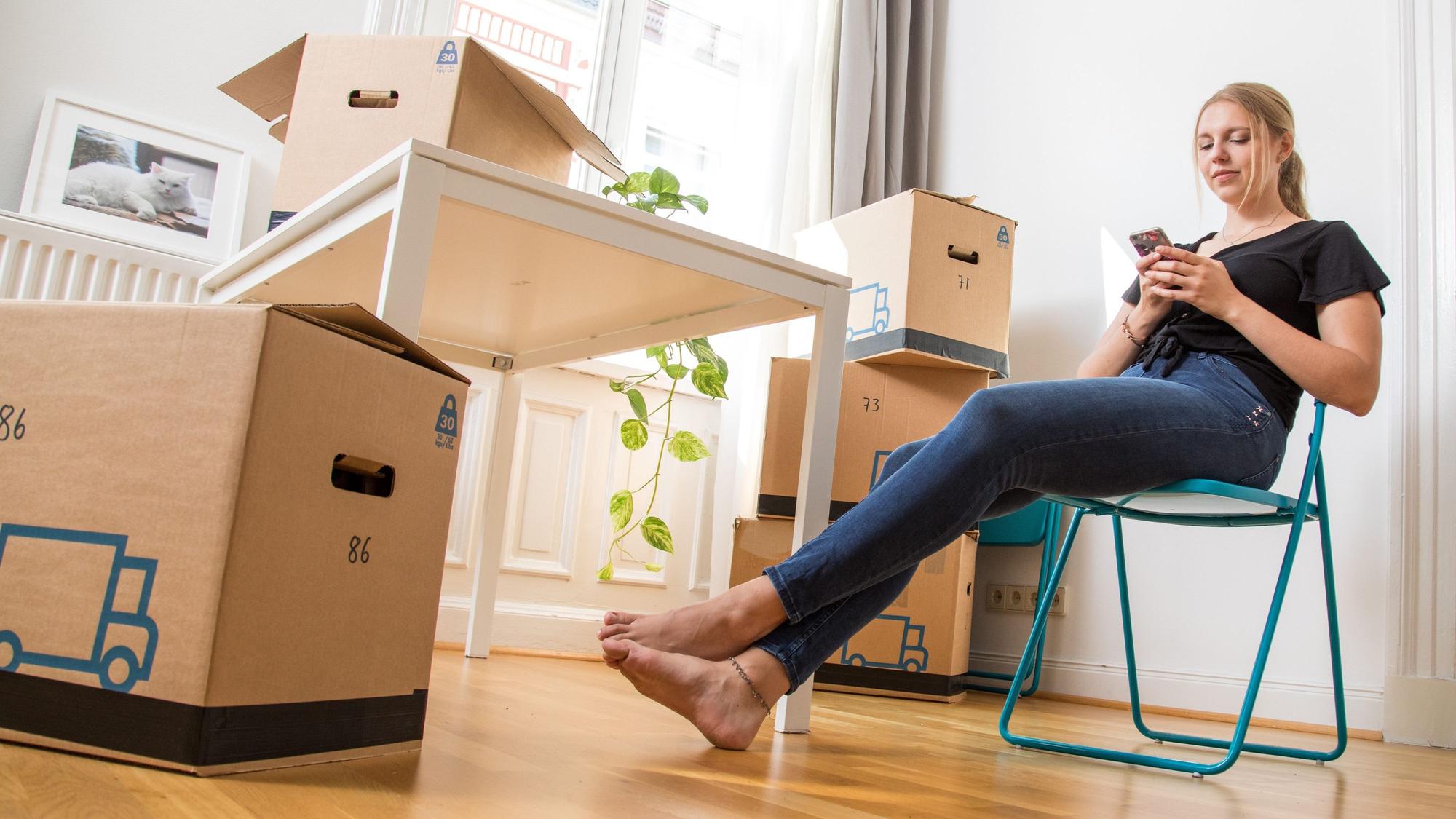 Eine Frau sitzt zwischen Umzugskartons in einer Wohnung auf einem Klappstuhl.